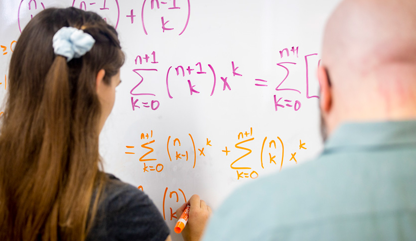 A mathematics student solving a mathematical equation on a whiteboard under the supervision of a mathematics faculty member.